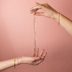 Hands holding a gold filled paperclip chain necklace with elongated links against a soft pink background.