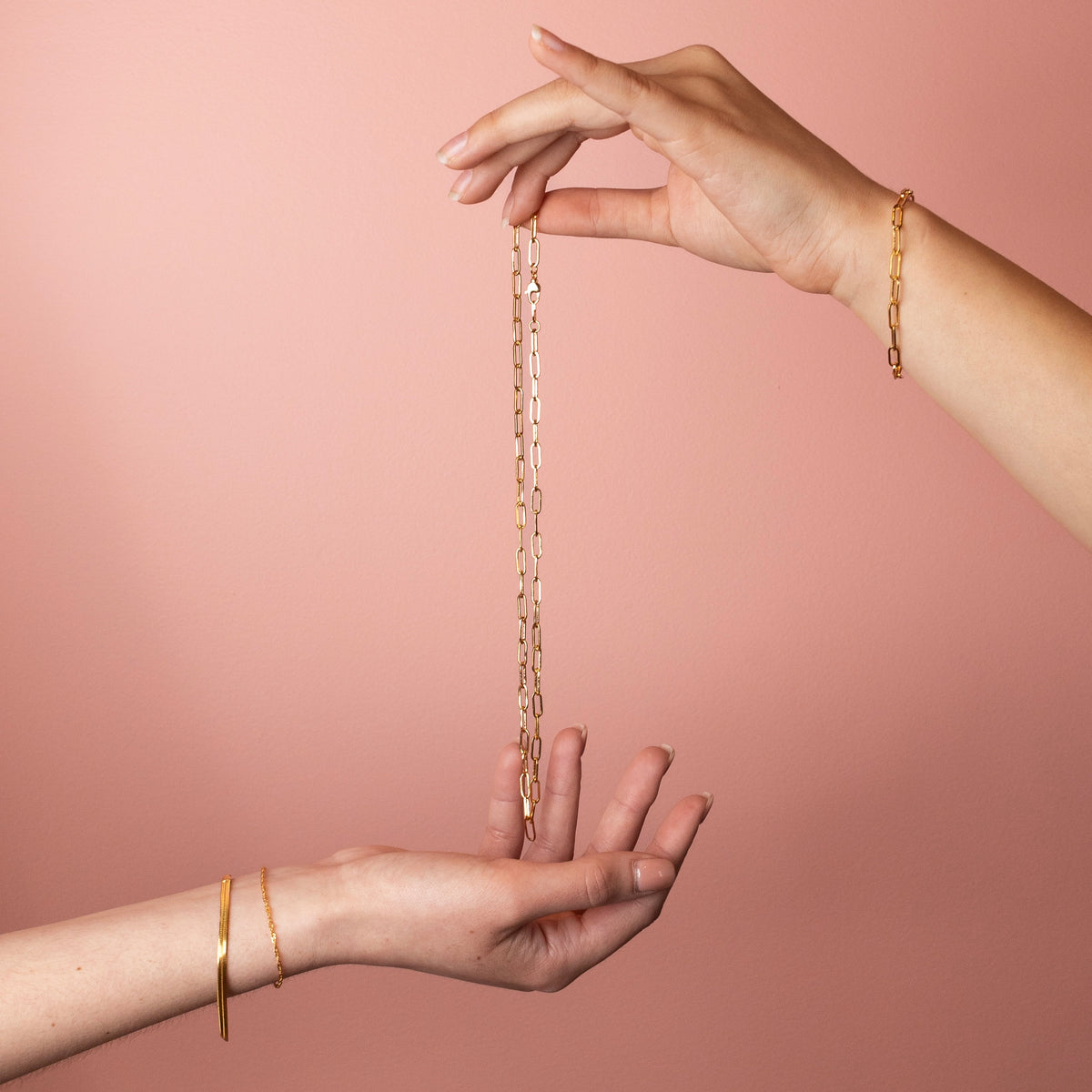 Hands holding a gold filled paperclip chain necklace with elongated links against a soft pink background.