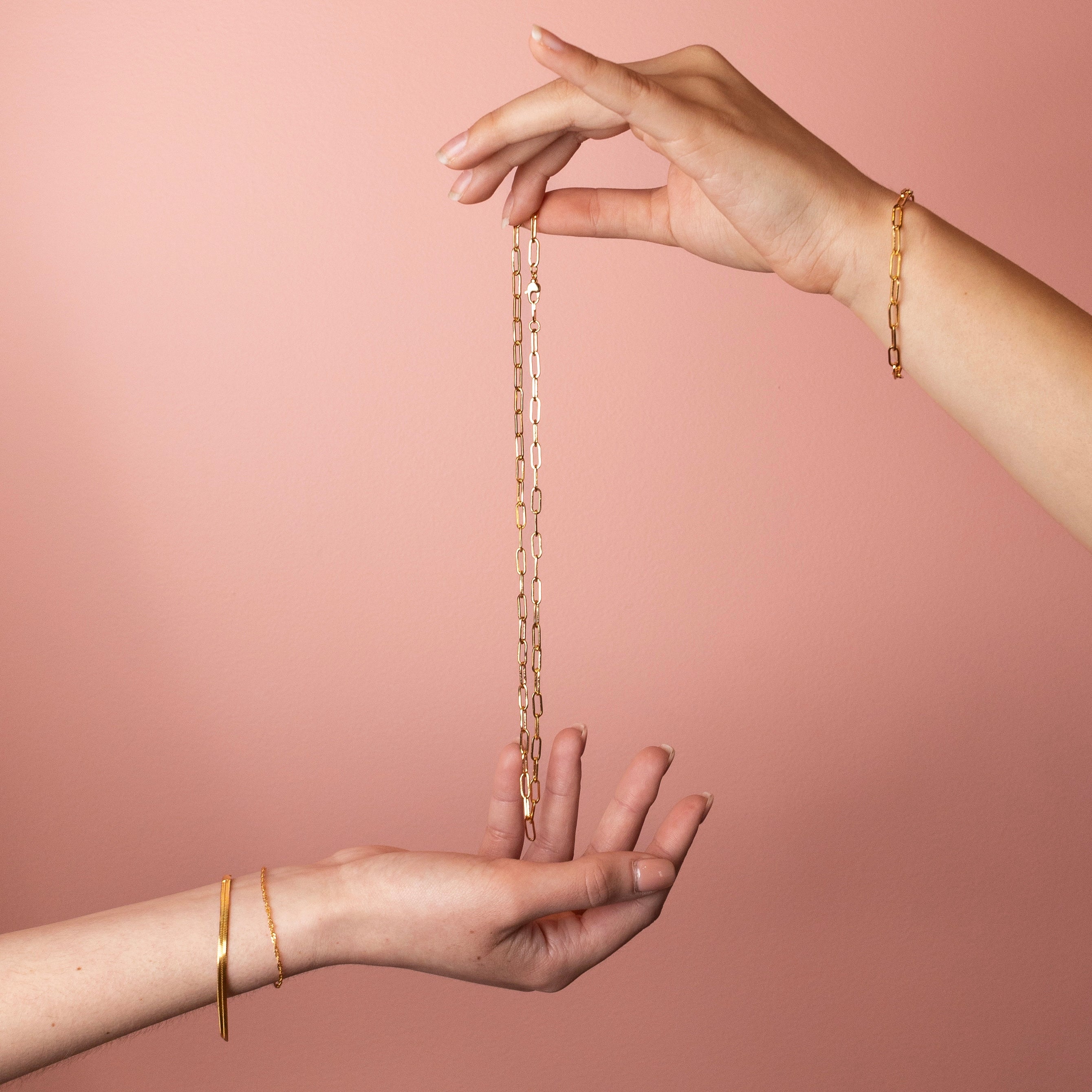 Hands holding an 18K gold fill paperclip chain necklace with elongated links against a soft pink background.
