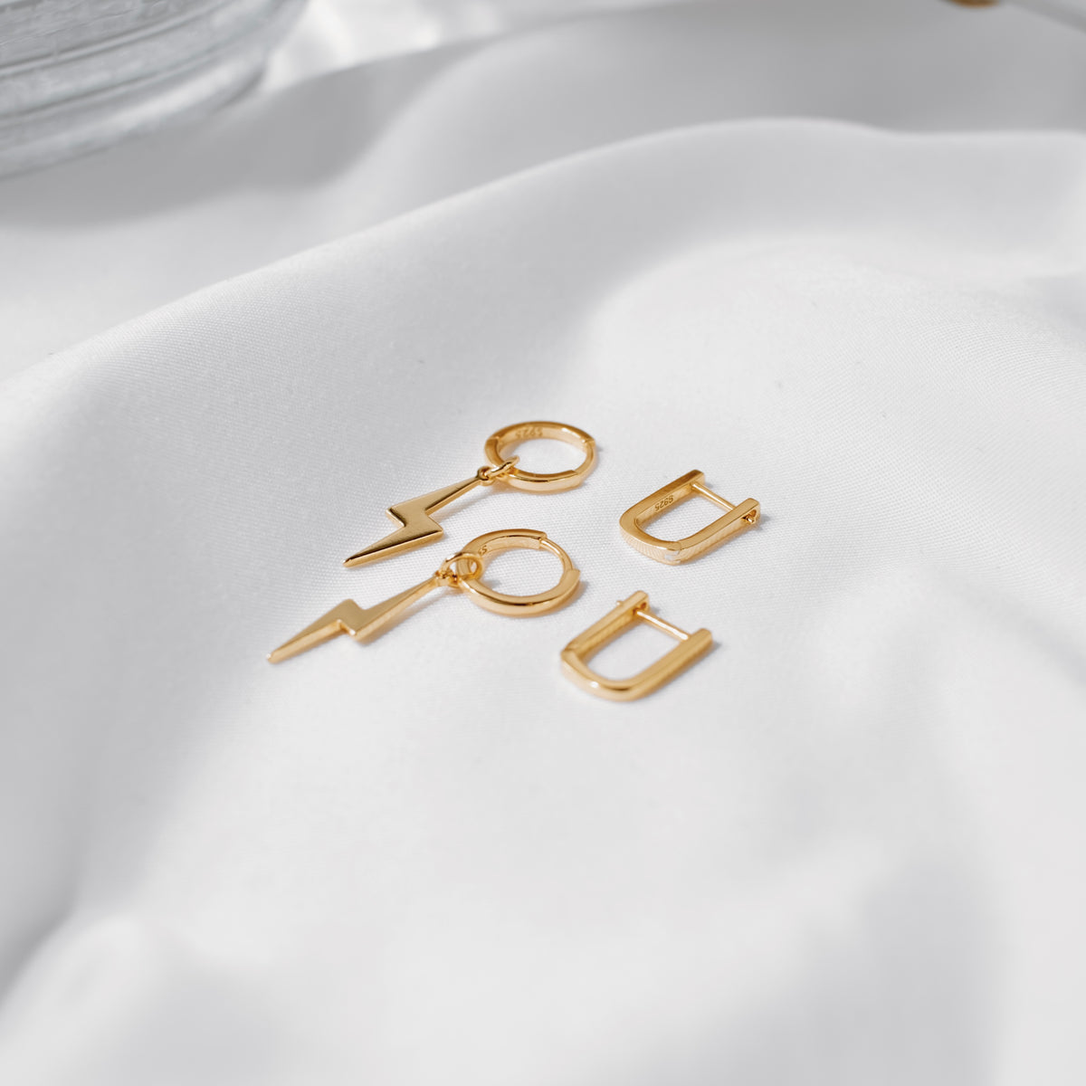 Gold vermeil oval huggie hoops shown with lightning-bolt charm earrings on white silk.