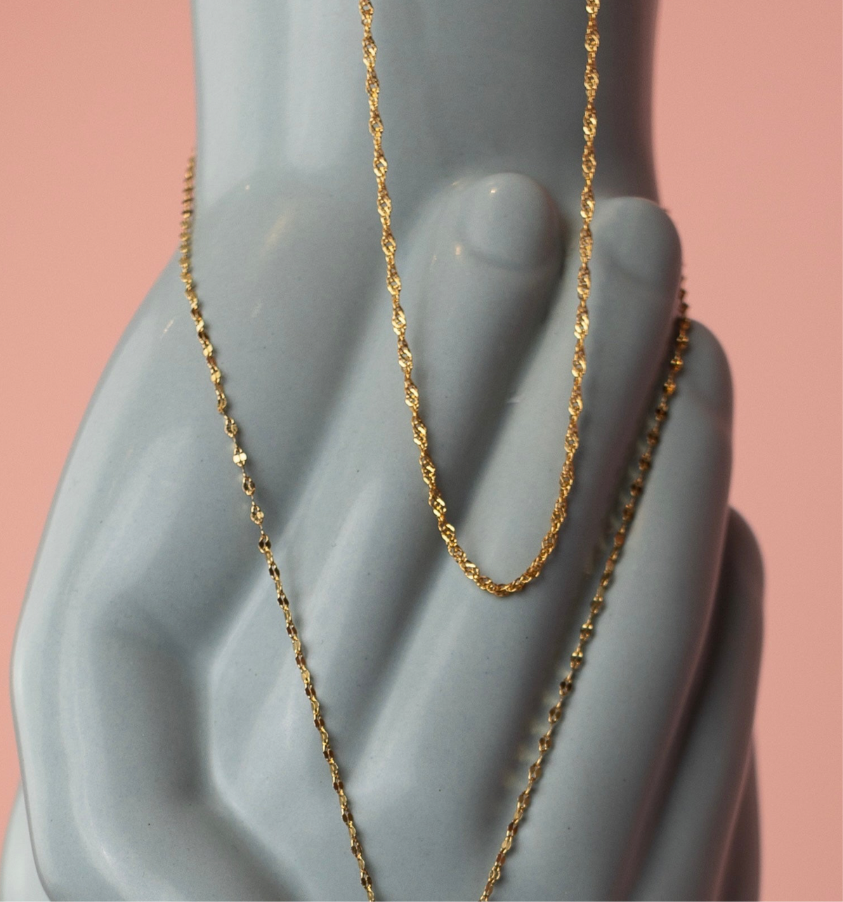 Gold filled Wave chain with twisted detailing draped over a ceramic hand, pink background.