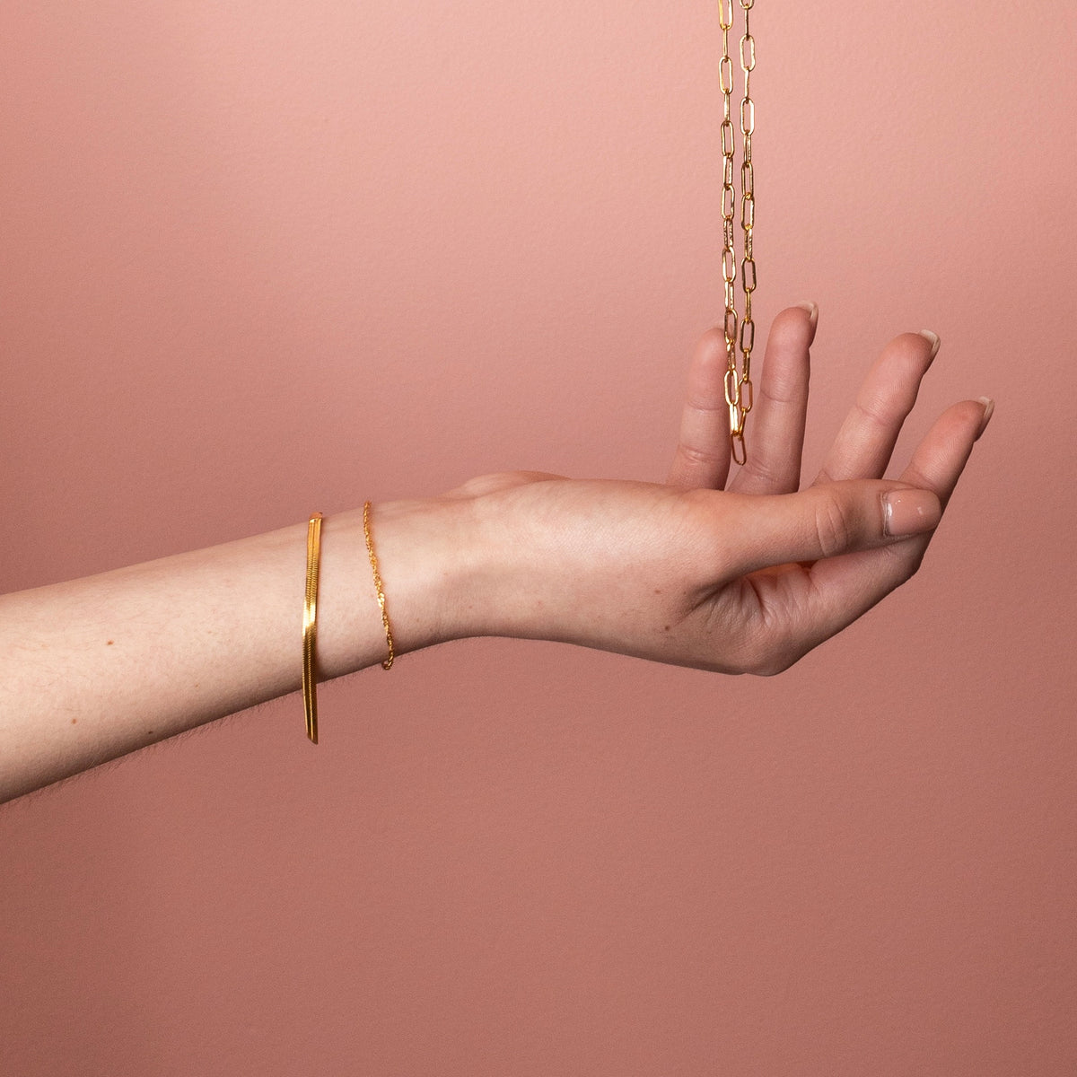 Gold plated stainless steel herringbone bracelet on a model's wrist against a soft pink background.