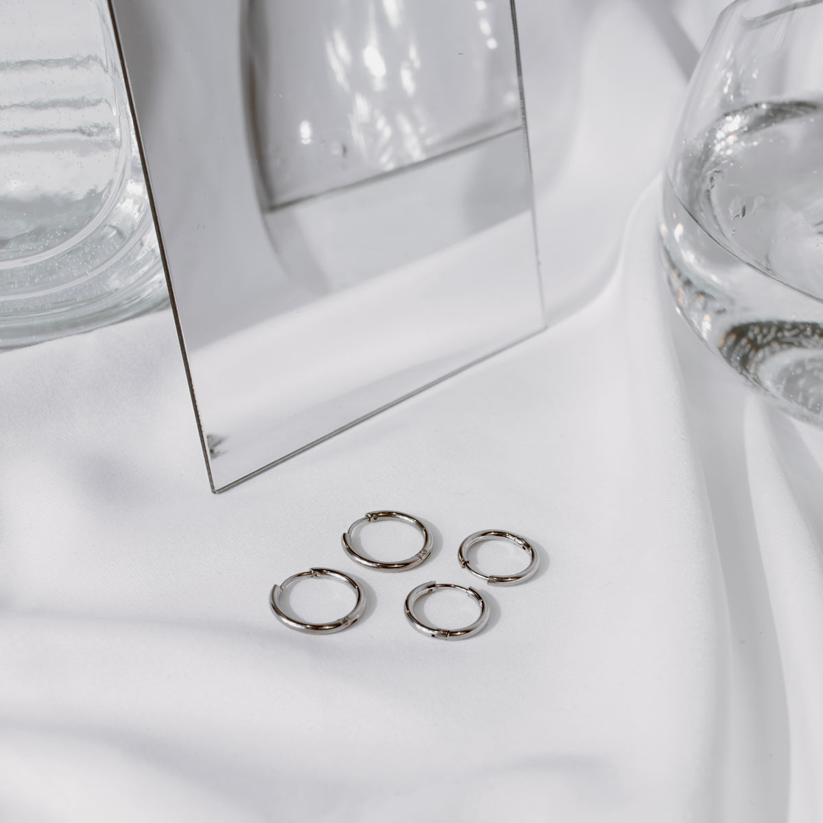 Four silver-tone stainless steel classic hoops clustered on white fabric beside a mirror.