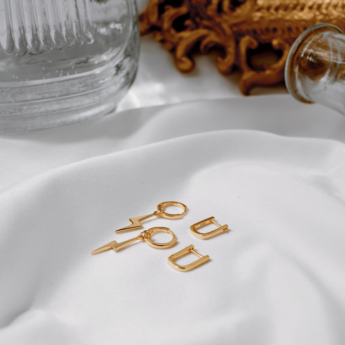 Gold vermeil oval huggie hoops with lightning-bolt charm earrings on white beside an ornate gold frame.