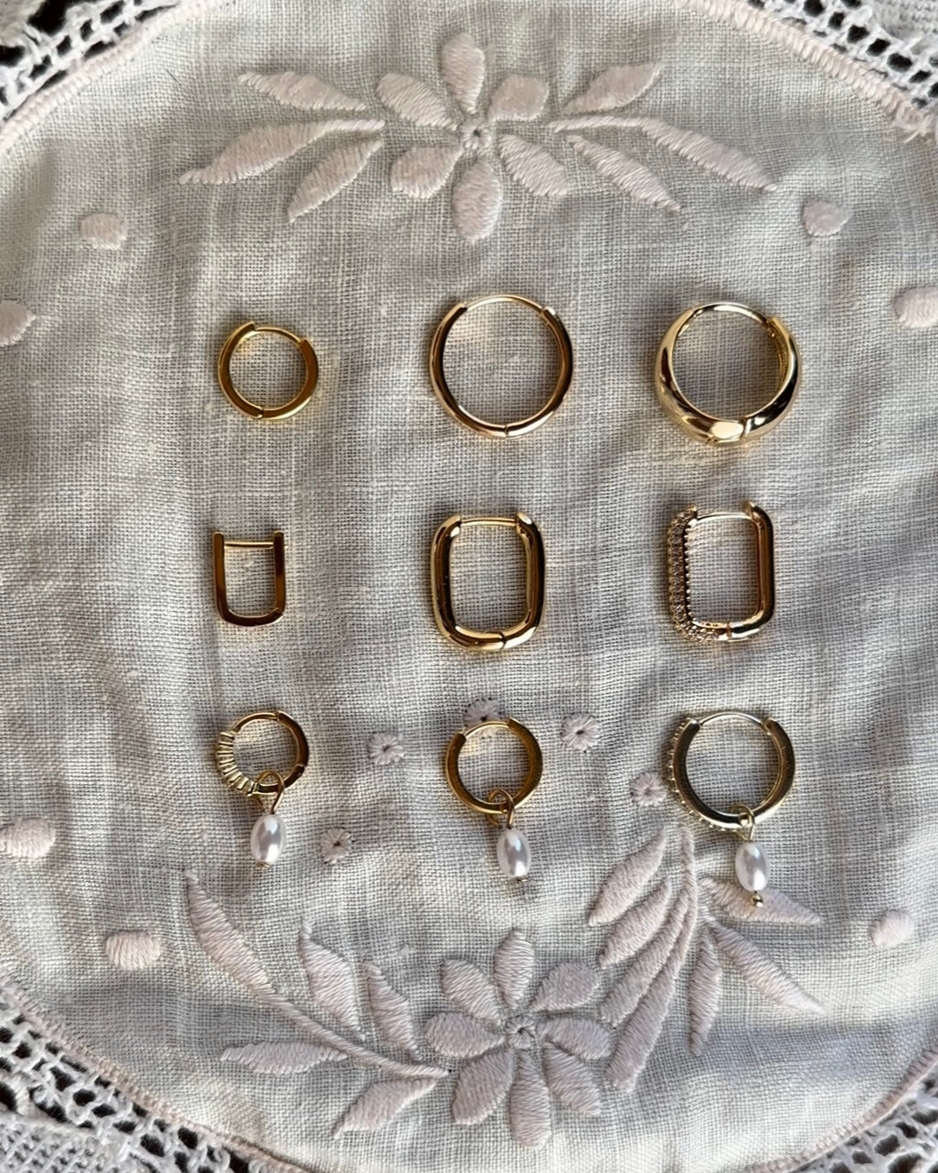 Flat-lay of gold and silver huggie hoops in several styles on embroidered linen.