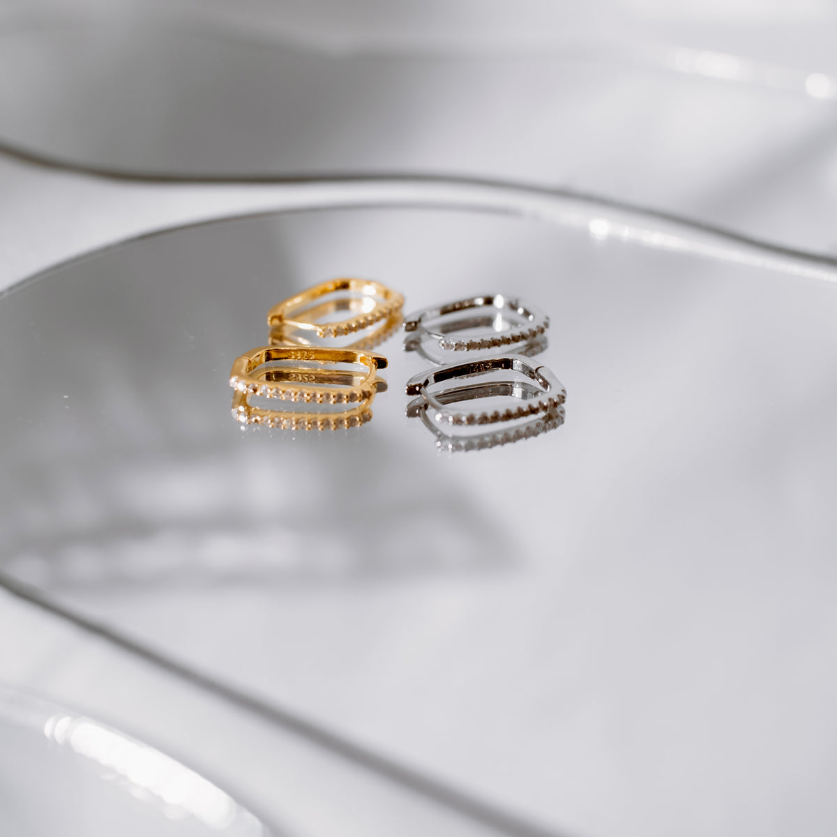 Gold vermeil and sterling silver oval huggie hoops on a mirrored surface.