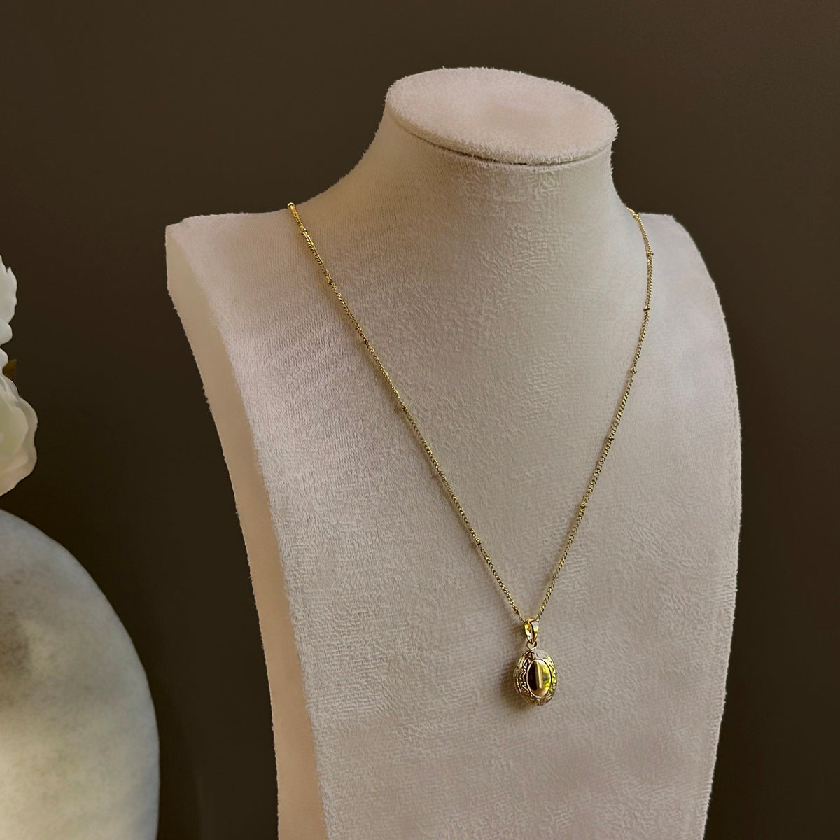 Oval locket on a satellite chain, shown on an ivory bust beside a white vase.