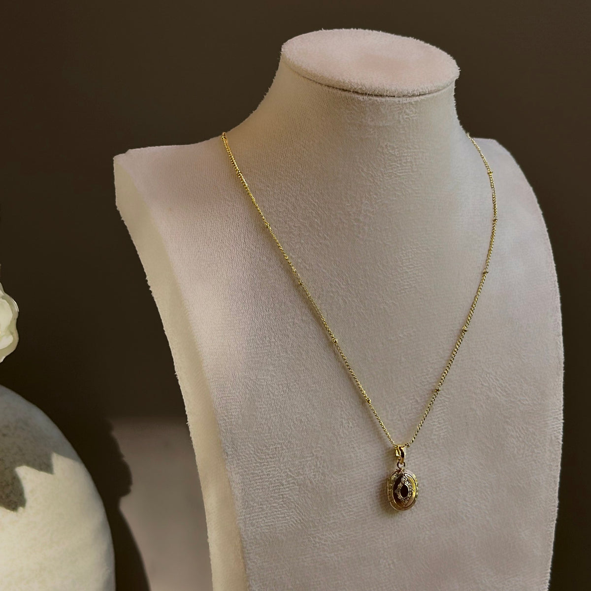 Small Ornate Oval locket on a satellite chain, shown on an ivory bust beside a white vase.