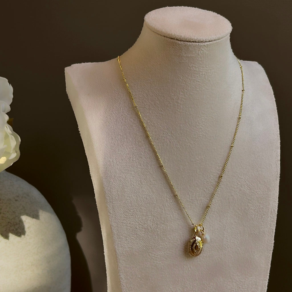Small Ornate Oval locket with its pearl charm, on a satellite chain on an ivory bust.