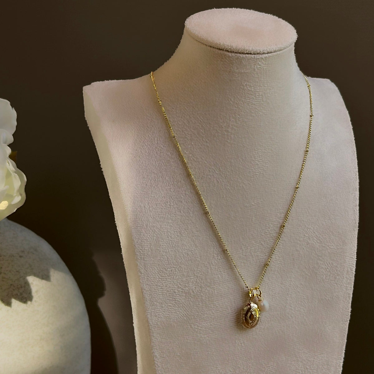 Small Ornate Oval locket with its pearl charm, on a satellite chain on an ivory bust.